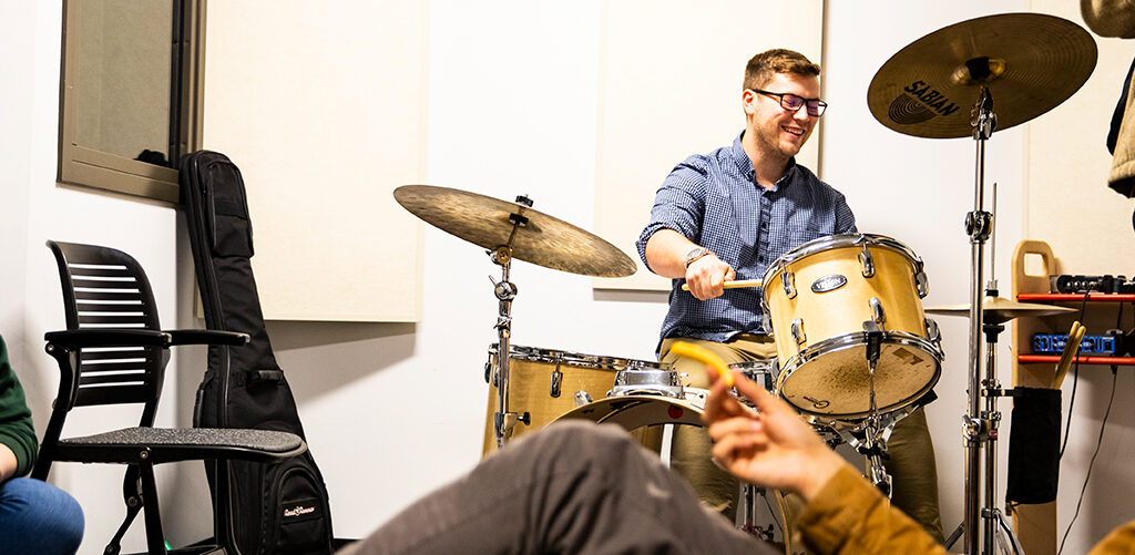 Student playing drums with other students laughing around him